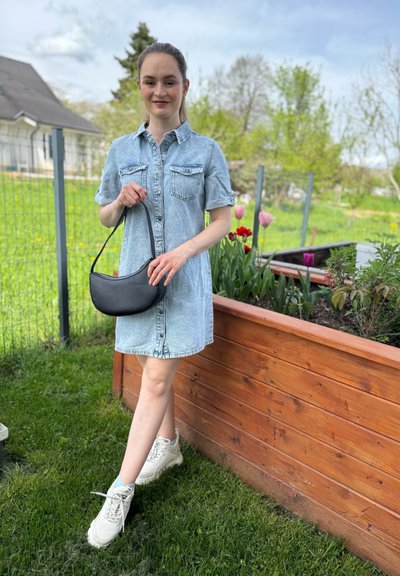 Vestido camisa de mezclilla de manga corta, color azul claro, con dos bolsillos en el pecho, combinado con un bolso negro de hombro curvado y zapatillas blancas.