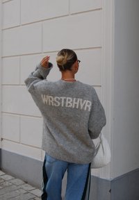 Grått strikket genser med hvit "WRSTBHR" tekst, oversized passform, ribbede mansjetter. Kombinert med lyseblå jeans med grønne sidestropper.