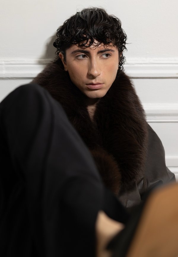 Col col en fourrure marron sur une veste en cuir foncé, portée sur une tenue noire. Cheveux bouclés foncés et expression faciale neutre sur un fond clair.