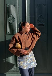 Kvinde i brun jakke og mønstret nederdel, der skygger for øjnene mod sollyset, holder en kaffekop og solbriller, stående foran en m ørkegrøn, udsmykket dør.