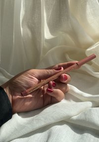 MAC leppeblyant i myk rosa med en teksturert, gylden mønstrede skaft. Holdt mot myk, hvit drapering. Hender med røde negler.