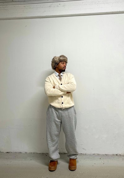 Cárdigan crema con botones negros, pantalones de chándal gris holgados, botas marrones y un sombrero de piel, de pie frente a una pared blanca lisa.