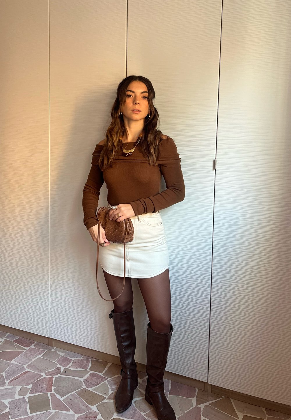 Hnědý top s dlouhým rukávem a zdobením na ramenou, bílá mini sukně, hnědá pletená kabelka, hnědé kozačky sahající ke kolenům, vrstvené náhrdelníky a průsvitné punčocháče.