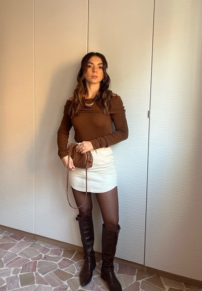 Top marrón de manga larga con detalles en los hombros, mini falda blanca, bolso tejido marrón, botas marrones hasta la rodilla, collares en capas y medias transparentes.