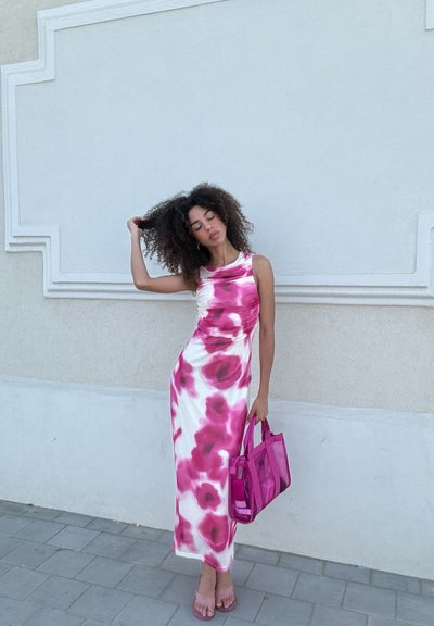 Vestido maxi de satén con un patrón floral en rosa y blanco, escote en forma de barco y drapeado en la parte delantera. Combinado con un bolso transparente rosa y sandalias.