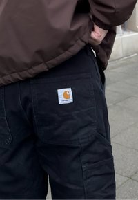 Άτομο που φοράει μαύρο παντελόνι Carhartt με ορατό λογότυπο στην πίσω τσέπη, ντυμένο με καφέ μπουφάν, που στέκεται σε πλακόστρωτη επιφάνεια σε εξωτερικό χώρο.