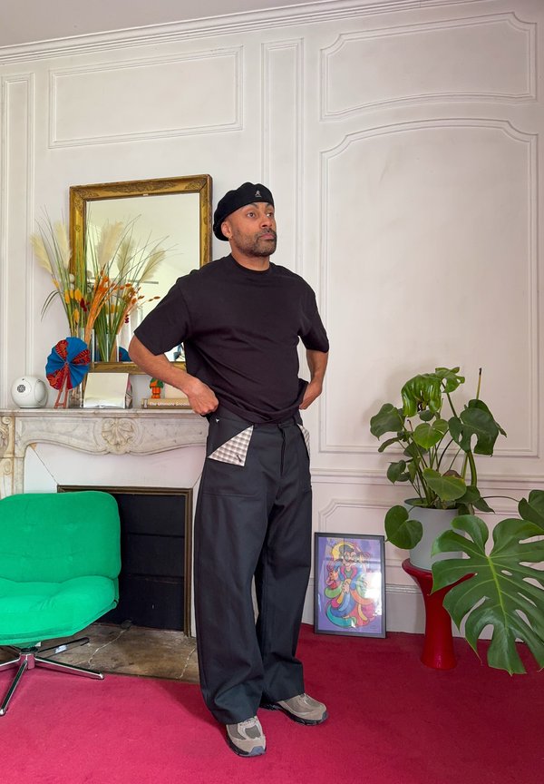 Homme en béret noir et pantalon se tient dans une pièce avec tapis rouge, chaise verte, plante en pot, œuvre d'art encadrée colorée et décoration sur la cheminée de chevet.