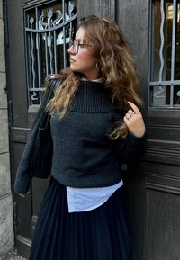 Μαύρο ριμπ πουλόβερ με ψηλό γιακά, φορεμένο πάνω από λευκό πουκάμισο. Συνδυασμένο με πλισέ μαύρη φούστα. Διακοσμημένο με δαχτυλίδια και κρεμαστά σκουλαρίκια.