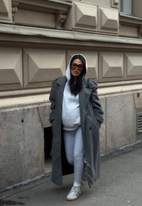 Femme portant un manteau gris surdimensionné par-dessus un sweat à capuche gris clair et des leggings, marchant sur un trottoir en ville les mains dans les poches du manteau.