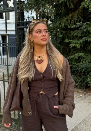 Brown knit cardigan, brown button-up top, and matching pants. Notable jewelry includes a large circular pendant and spiral earrings.
