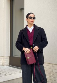 Abrigo de lana negro sobre un suéter de punto burdeos y una camisa blanca con cuello. Pantalones de pierna ancha grises, cinturón negro con detalles dorados y bolso burdeos.