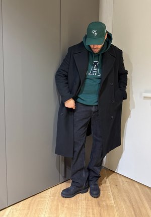 Hombre con abrigo largo oscuro, sudadera con capucha verde con la letra "A", gorra verde de NY, jeans oscuros y botas negras, de pie en el interior sobre un suelo de madera.
