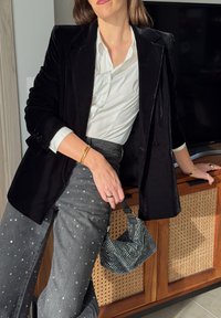 Crni somotski sako preko bijele košulje, uparen s sivim hlačama ukrašenim srebrnim zakovicama. Drži teksturiranu crnu torbu s ukrasima.