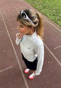 Kobieta w kucyku, ubrana w biały długi rękaw, czarne szorty, neonowe różowe i białe sneakersy oraz białe okulary przeciwsłoneczne na głowie, stojąca na torze.