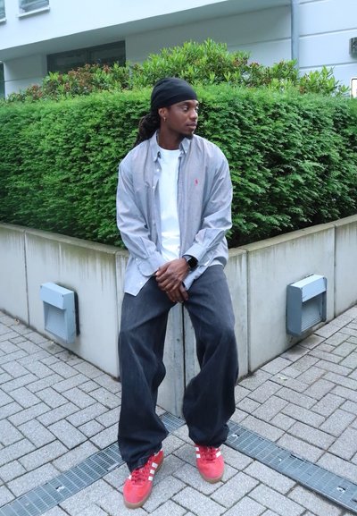 Camisa de botones gris sobre una camiseta blanca, jeans oscuros y holgados, y zapatillas rojas. Sentado cerca de un seto verde con una base de concreto.