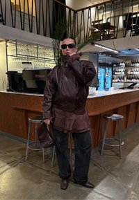 Ruskea nahkatakki, jossa korkea kaulus ja kerroksittainen muotoilu, yhdistetty tummiin farkkuihin ja vastaavaan käsilaukkuun, kahvilataustan edessä.