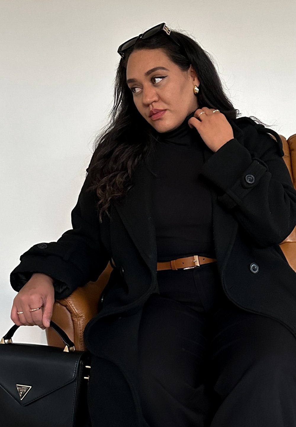 Mustat villakangastakki isot napit, poolopaita, ruskea nahkavyö, musta laukku kultaisilla yksityiskohdilla, istuu ruskealla nahkatuolilla.