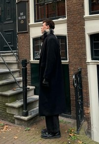 Μαύρο μακρύ παλτό, μαύρα γάντια και σκούρα παντελόνια φορεμένα από ένα άτομο που στέκεται δίπλα σε ένα κτίριο με καφέ το ύβλα που έχει μια περίτεχνη μαύρη πόρτα και σκαλιά.