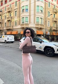 Jasnoróżowy, dziergany komplet dwuczęściowy z dopasowanym topem i rozszerzanymi spodniami. Brązowa skórzana torebka o usztywnionym kształcie. Miejski krajobraz z budynkami.