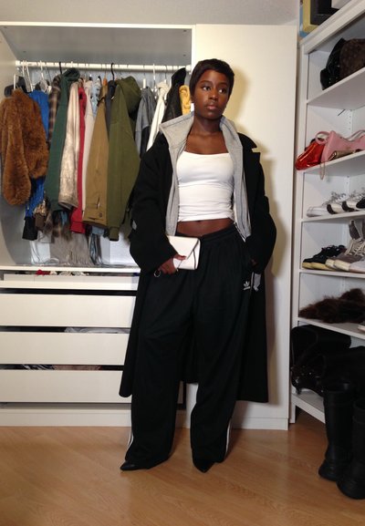 Mujer con un top corto blanco, pantalones anchos negros y chaquetas superpuestas está en un vestidor con ropa colgante y estantes de zapatos y bolsos.