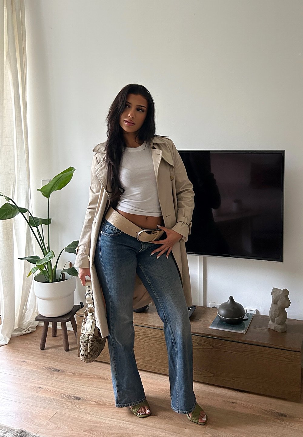 Beżowy trencz na biały top, w połączeniu z rozkloszowanymi niebieskimi dżinsami. Trzyma wzorzystą torebkę, stoi przed telewizorem i rośliną.