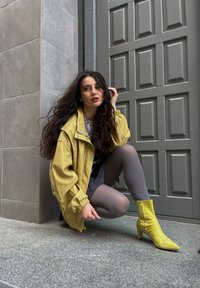 Gelbe übergroße Jacke, graues Oberteil, graue Strumpfhose und strukturierte gelbe Knöchelstiefel mit spitzer Zehe. Der Hintergrund zeigt eine strukturierte graue Wand.