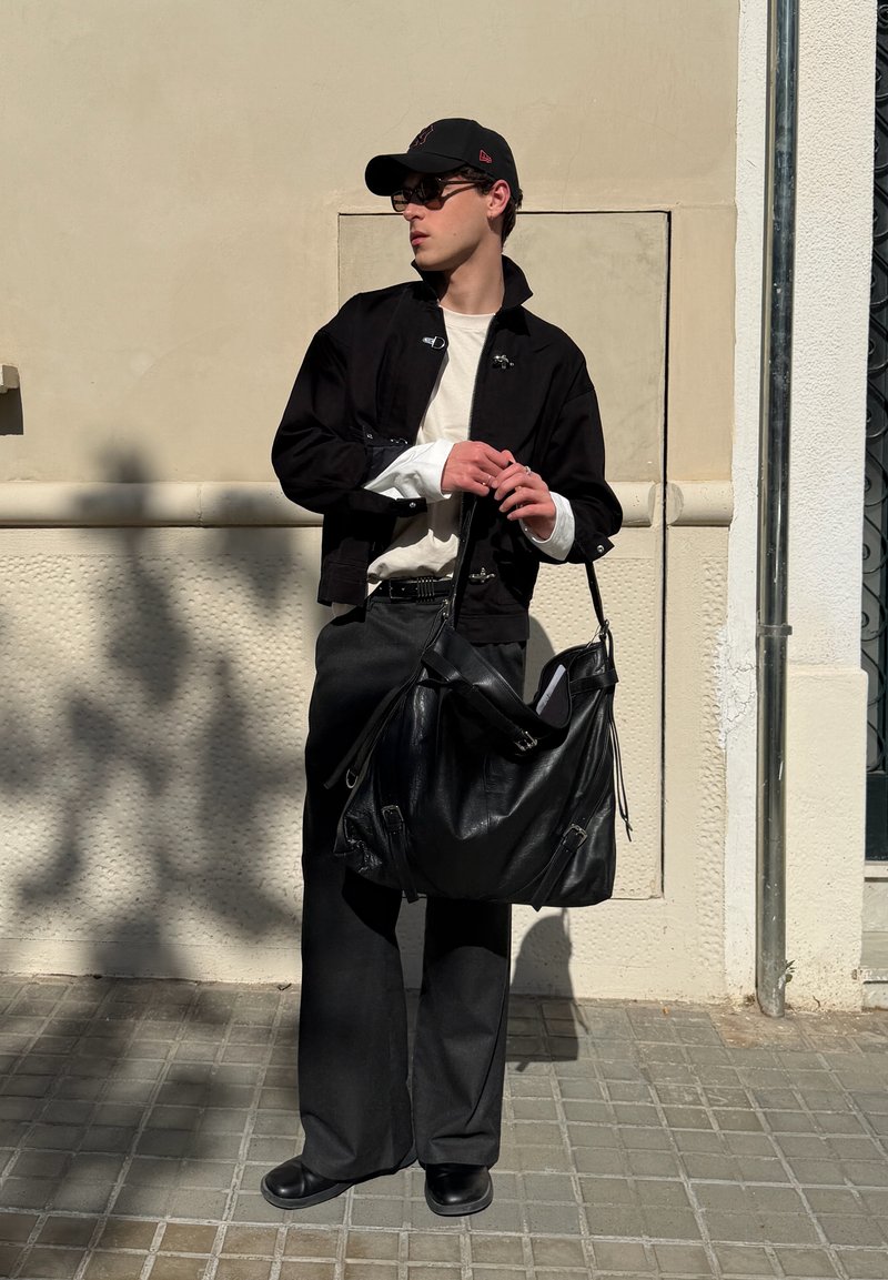 Jeune homme portant une casquette noire, des lunettes de soleil, une veste noire et un pantalon à jambes larges, debout sur un trottoir carrelé tenant un grand sac en cuir noir.