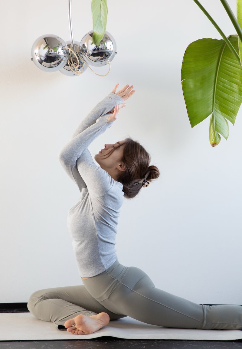 Femme vêtue de gris effectuant un étirement de yoga assis sur un tapis, près de grandes feuilles vertes et d'une lampe de plafond ronde en chrome.