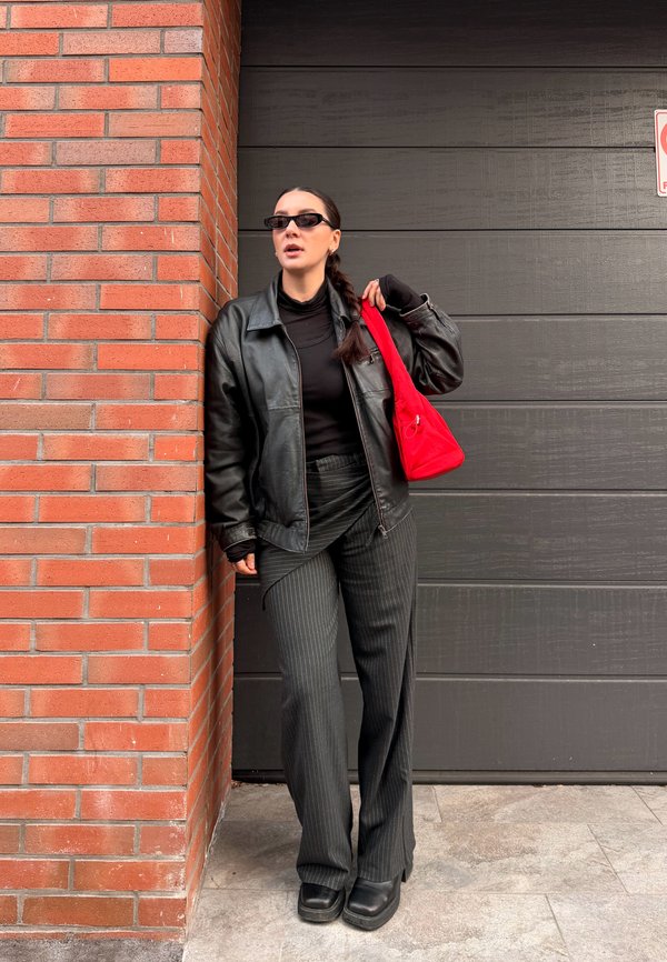 Frau trägt schwarze Lederjacke, Nadelstreifenhose, schwarze Sonnenbrille, hält eine rote Handtasche, steht an einer Backsteinmauer und vor einer dunklen Garagentür.