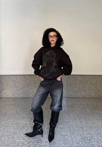 Svart overdimensjonert hettegenser med grafisk design, grå store jeans og knelengde sorte lakkboots. Modellen står mot en lys vegg.