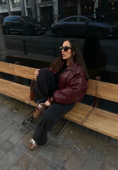 Chaqueta acolchada burdeos, top negro y pantalones anchos negros. Zapatos marrones con cordones blancos. Sentado en un banco de madera. Entorno urbano.