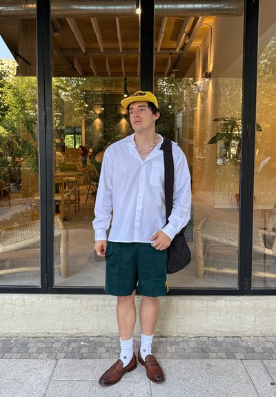 Camisa blanca de manga larga con botones, pantalones cortos verdes con acentos amarillos, mocasines marrones, bolso negro de hombro y gorra amarilla. Ambientación exterior brillante.
