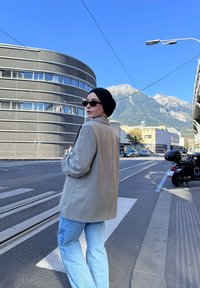 Blazer gris clair au design structuré, associé à un jean bleu décontracté. Un foulard noir et des lunettes de soleil complètent le look, sur fond urbain.