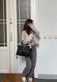 Bežna skrajšana jakna, siva majica brez rokavov, sive kavbojke in črna usnjena torba z resicami in kovančki. Srebrni športniki na rjavem preprog.