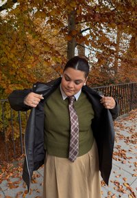 Svart skinnjakke over en grønn genser med ribbet kant, beige skjørt og rutet slips. Mot en bakgrunn av høstløv og nedfalne blader.