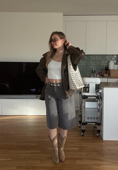 Chaqueta marrón de gran tamaño, top corto blanco, pantalones cortos gris de pierna ancha, botas de vaquero beige. Bolso con tachuelas plateadas. Suelo de madera clara.
