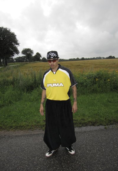 Camisa deportiva negra y amarilla con el logo de "PUMA", pantalones anchos negros, gorra negra con letras blancas y gafas de sol, de pie en una carretera.