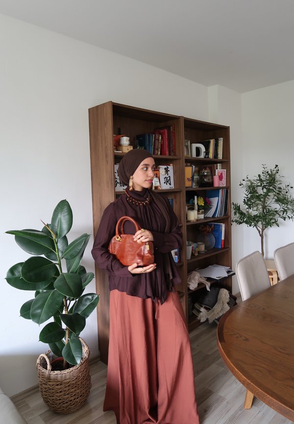 Barna hijabos, rozsdabarna szoknyás nő bézs táskát tart, és egy könyvespolc és növények melletti világos szobában áll, ahol fa asztal és székek vannak.