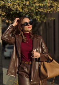 Blazer y pantalones de cuero sintético marrón combinados con un suéter de cuello alto de canalé burdeos. Los accesorios incluyen gafas de sol negras y un bolso tipo cubo color beige.