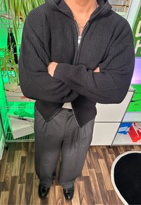Schwarze, gerippte Zip-Hoodie mit Kapuze, kombiniert mit grauen, plissierten Stoffhosen und schwarzen Schuhen. Der Hintergrund zeigt Pflanzen und bunten Stauraum.