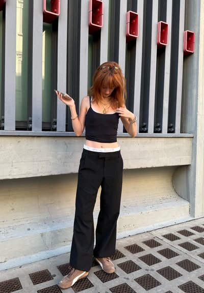 Top sin mangas negro, pantalones negros de tiro alto y zapatillas marrones. Collares de cadena notables y texturas en capas. Fondo gris con acentos rojos.