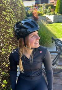 Casco de ciclismo negro con ranuras de ventilación, llevado por una persona en un traje de ciclismo negro de manga larga con un logo, junto a una bicicleta.