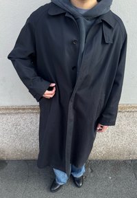 Άτομο που φοράει μακρύ μαύρο παλτό πάνω από σκούρα φούτερ με κουκούλα, μπλε τζιν και μαύρα παπούτσια, στέκεται σε πλακόστρωτο έδαφος κοντά σε πέτρινο τοίχο.