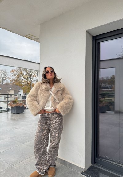 Chaqueta de piel sintética beige, top de punto blanco y pantalones gris con estampado de leopardo. Los accesorios incluyen gafas de sol redondas y botines marrones. Ambiente interior/exterior.