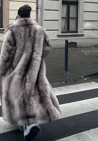 Sivý kabát z umelej kožušiny s dlhým, nadýchaným dizajnom a širokým golierom, zdobený tmavšími vertikálnymi pruhmi a plynúcou textúrou.