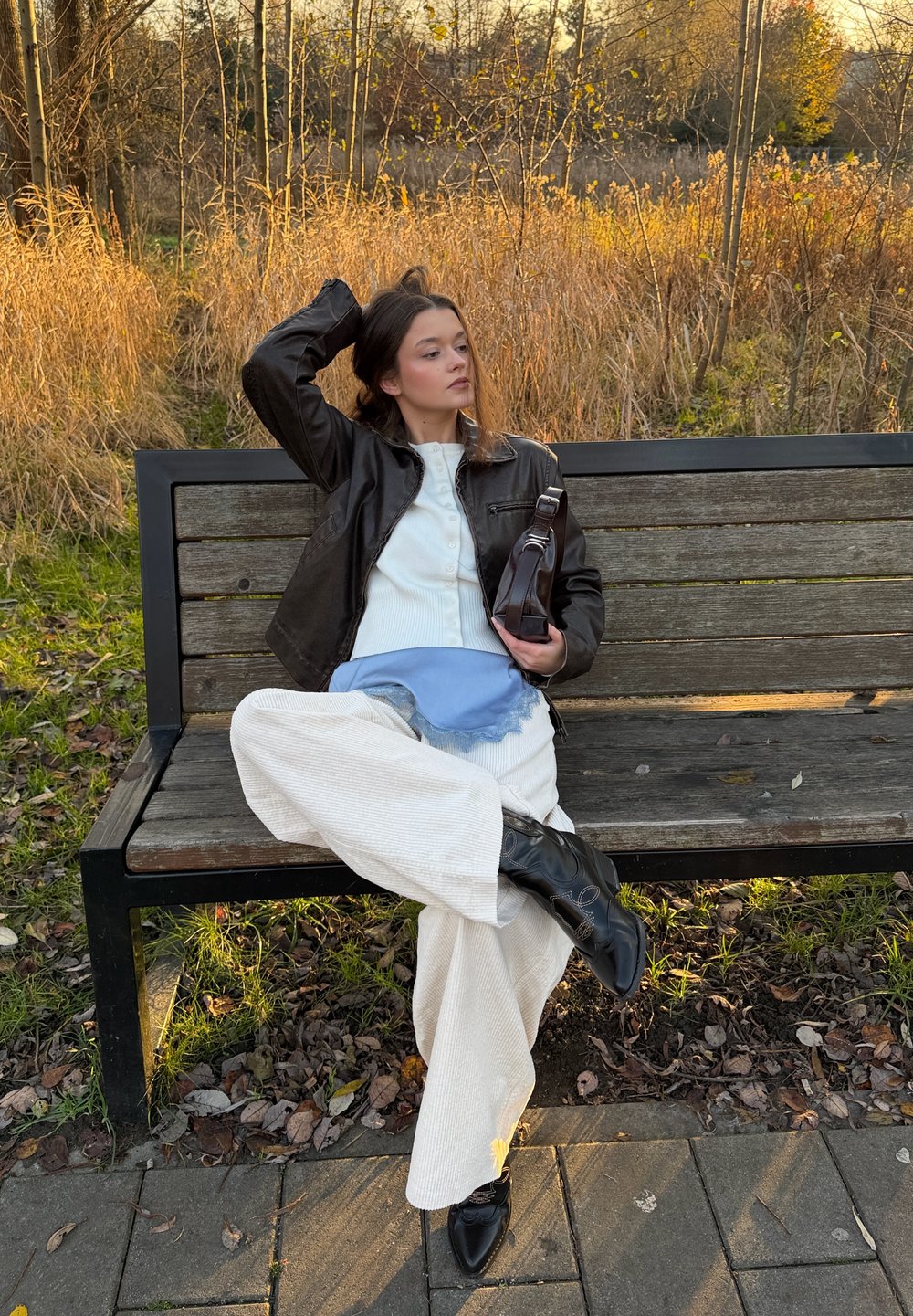 Giacca di pelle nera, top bianco a coste, sciarpa azzurro chiaro con frange, pantaloni ampi color crema, stivali neri al ginocchio. Seduta su una panchina di legno.