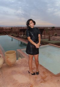 Czarna sukienka koszulowa ze skóry z krótkimi rękawami, w zestawie z czarnymi szpilkami i trzymająca czarną torebkę o fakturze, stojąca przy basenie.