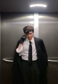 Manteau noir, chemise blanche, cravate noire et pantalon à fines rayures. Porte des gants noirs et des lunettes, debout dans un ascenseur aux parois métalliques.