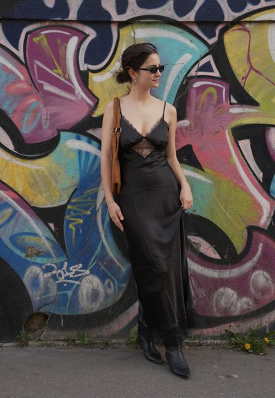 Mujer vestida con un vestido de satén negro y gafas de sol, de pie junto a una pared de grafiti colorida, sosteniendo un bolso marrón de hombro y posando con una postura relajada.