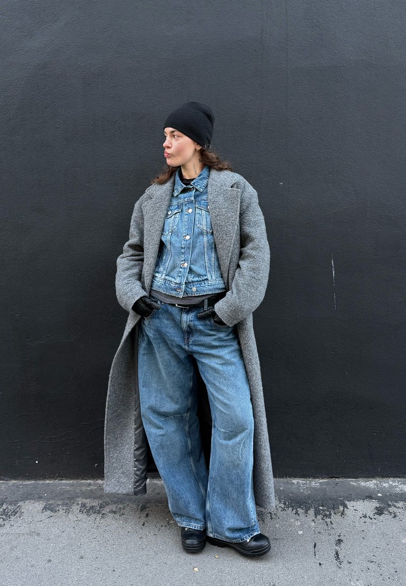 Personne portant un bonnet noir, un long manteau gris par-dessus une veste en denim et un jean à jambes larges, debout les mains dans les poches contre un mur sombre.
